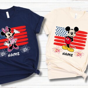 May include: Two t-shirts featuring cartoon characters in front of an American flag design. One shirt is navy blue with Minnie Mouse, the other is cream-colored with Mickey Mouse. Both shirts have the word "NAME" printed below the characters.