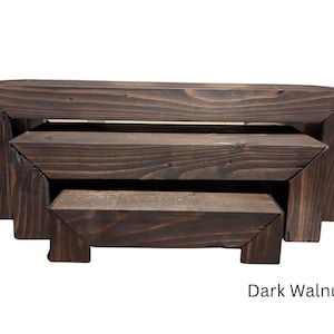 May include: A set of three dark walnut wooden display risers. The risers are rectangular with a stepped design, each level slightly smaller than the one below. The wood grain is visible, and the finish is a deep brown.