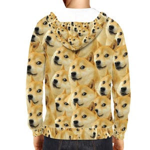 Puede incluir: Una sudadera con capucha con un patrón repetido de caras de memes Doge. El diseño cubre toda la espalda y las mangas de la prenda. Las caras de Doge son en tonos beige y marrón, con ojos y narices negros.