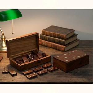 Peut inclure: Un jeu de dominos en bois avec une boîte, comprenant des dominos marron foncé avec des points blancs. L'ensemble est présenté sur une table en bois, à côté d'une boîte en bois fermée, de livres et d'une lampe de bureau à abat-jour vert. Les dominos sont éparpillés.