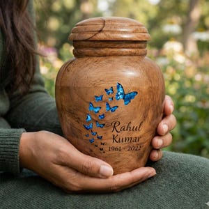 Puede incluir: Urna de madera con tapa, adornada con mariposas azules. La urna está grabada con el nombre "Rahul Kumar" y los años "1961-2022". La urna está sostenida en las manos.
