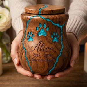 May include: A wooden urn with a natural brown wood grain finish, featuring a decorative turquoise resin inlay that runs across the lid and body. The urn is engraved with the name Alice, the years 2012-2019, the phrase forever loved, and paw prints.