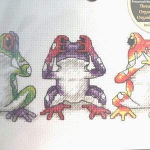 May include: Cross-stitch pattern featuring three colorful frogs in a 'see no evil, hear no evil, speak no evil' pose. The frogs are green, purple, and orange, with white bellies and red feet. The pattern is on a white background.