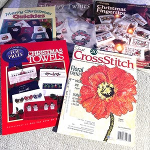 May include: A collection of cross-stitch pattern books and magazines. Titles include "Christmas Towels," "Merry Christmas Quickies," and "Just CrossStitch." The magazines feature floral designs and holiday-themed patterns.