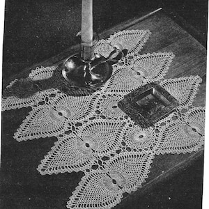 May include: A vintage, off-white crocheted doily with a repeating geometric pattern. The doily is placed on a dark wooden surface, with a silver candle holder and a small square dish. The doily's design features circular and leaf-like shapes.