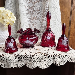 May include: A collection of ruby red glass decorative items, including two bells and a small vase, each adorned with white floral designs. The items are arranged on a crocheted lace surface, creating a vintage aesthetic.