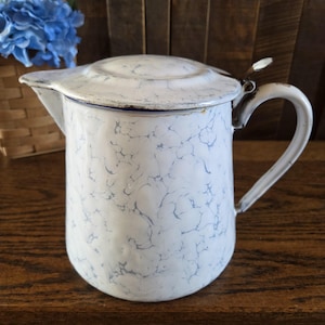 May include: A white enamel pitcher featuring a light blue marbled pattern throughout the exterior. The pitcher includes a matching lid with a metal hinge, a curved handle on the side, and a small pouring spout. The finish shows signs of wear.