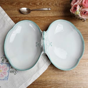 May include: A white ceramic serving dish with a unique double-bowl design. The dish has a light teal trim and decorative accents. A silver spoon rests nearby on a wooden surface, with a floral arrangement in the background. This elegant dish is perfect for serving appetizers.