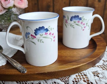 May include: Two white ceramic mugs with blue floral designs and blue rims sit on a wooden tray. The mugs have curved handles and are decorated with blue and pink flowers and green leaves. A spoon and a vase of pink flowers are in the background.