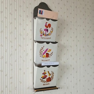 May include: A vertical wall-mounted metal organizer with three cream-colored pockets. Each pocket features brown text and colorful illustrations: a rooster for LETTERS, a rolling pin for MEMOS, and a tiered tray for MISC. A pencil rests at the base.