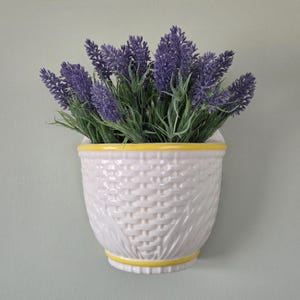 May include: A white ceramic wall planter with a textured basket-weave pattern and yellow trim, filled with a dense arrangement of artificial purple lavender sprigs with green stems, mounted against a solid light gray wall surface.