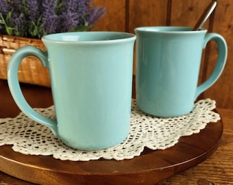 May include: Two light blue ceramic mugs with curved handles sit on a wooden tray with a white lace doily. A basket of purple flowers is in the background. One mug has a spoon inside.