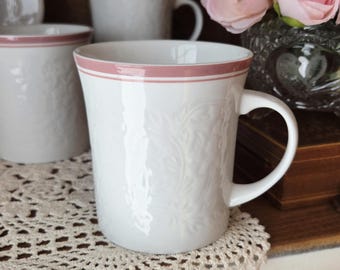 May include: White ceramic mug with a pink rim and a floral embossed design. The mug has a curved handle and is sitting on a lace doily. Additional mugs are visible in the background.