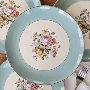 May include: A collection of vintage ceramic plates. Each plate features a light blue rim, a cream-colored center, and a floral bouquet design in shades of pink, yellow, and brown. A silver fork rests on a white lace doily.
