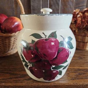 May include: A cream-colored ceramic canister with a lid, featuring a painted design of deep red apples and green leaves. The rim of the canister has a distressed blue-grey finish, and the body has a rounded shape with a smooth, glossy surface.