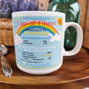May include: White ceramic mug with the text "Cup of Cheer" and a rainbow design. The mug includes nutritional information for faith and optimism, and a guarantee. The mug is on a wooden tray.