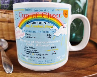 May include: White ceramic mug with the text "Cup of Cheer" and a rainbow design. The mug includes nutritional information for faith and optimism, and a guarantee. The mug is on a wooden tray.