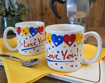 May include: Two white ceramic mugs with the text "I Love You!" and colorful heart and confetti designs. A silver spoon rests on a yellow napkin, with a coffee maker in the background.