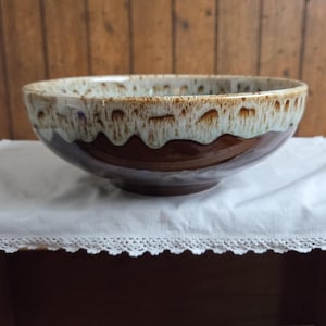 May include: A round ceramic bowl featuring a two-tone glaze. The upper portion has a cream-colored glaze with brown drip patterns, while the lower portion is finished in a solid, glossy dark brown glaze. The bowl has a wide, open rim.