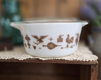 Op de afbeelding: Witte Pyrex ovenschaal met een doorzichtig deksel. De schaal heeft bruine decoratieve afbeeldingen van een adelaar, een haan en andere vintage keukenthema's. De schaal staat op een wit kanten kleedje.