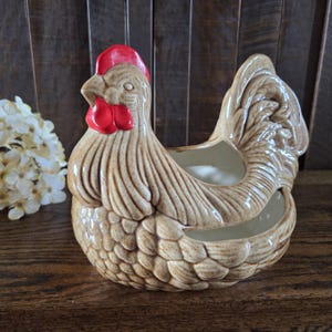 May include: A ceramic chicken-shaped basket or planter with a speckled tan finish and a bright red comb and wattle. The piece features molded feather details across the body and wings, with a hollow center and an open top for storage or display.