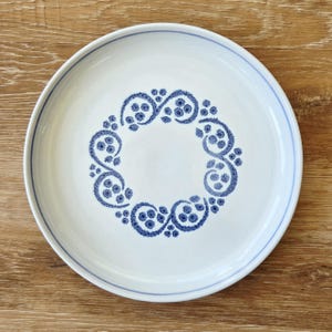 May include: White ceramic plate with a blue floral design around the center. The plate has a blue border and a decorative blue pattern. The plate is on a wooden surface.