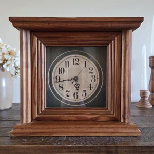 May include: A square wooden mantel clock with a medium brown finish and a stepped frame design. The clock face is circular with a cream background, black Arabic numerals, and ornate black metal hands indicating the time at approximately 8:25.