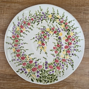 May include: A white ceramic plate with a hand-painted floral design. The design features a wreath of red roses, purple and yellow flowers, and green foliage. The plate is round and has a glossy finish.
