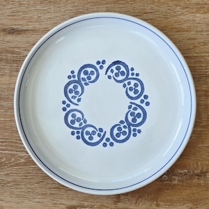 May include: White ceramic plate with a blue floral design in the center and a blue border. The plate has a circular shape and is set against a wooden surface.