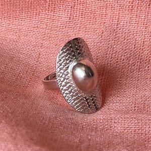 May include: Silver-toned ring with a textured, oval-shaped face and a raised, rounded center. The ring's surface has a hammered appearance, and it sits on a pink fabric background. The ring is a piece of jewelry.