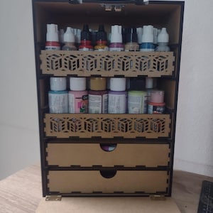 Puede incluir: Una caja de almacenamiento de pintura de madera con un asa negra y recortes decorativos. La caja tiene múltiples estantes y cajones llenos de varios botes de pintura. Diseñada para organizar y almacenar suministros de arte.