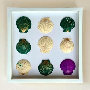May include: A square white frame containing nine scallop shells arranged in a three-by-three grid. The shells feature various colors including deep green, cream, white, and purple, with some shells decorated with gold-colored line accents.