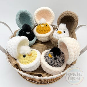 May include: A wicker basket holds six crocheted chicks emerging from fluffy eggshells. The chicks are various colors, including yellow, white, black, and gray, with fluffy accents. The eggshells are in shades of white, beige, and blue. The basket sits on a burlap surface.
