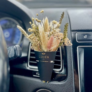 May include: Dried flower bouquet in a black paper cone with the word "LUCCA" and the website "www.luccabirthday.com" printed on it. The bouquet is placed in a car's air vent.