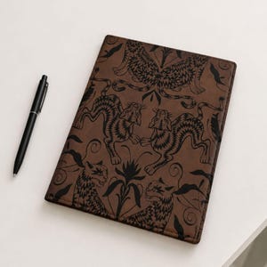 May include: A rectangular brown notebook featuring a repeating black graphic print of stylized tigers and floral elements, accompanied by a slim black ballpoint pen with a silver clip positioned to the left of the notebook cover.