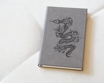 Könnte beinhalten: Ein graues, ledergebundenes Tagebuch mit einer schwarz-weißen Illustration einer Schlange, die mit Rosen verschlungen ist.