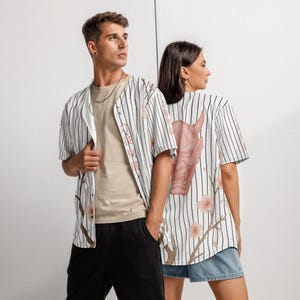 Op de afbeelding: Baseballshirt met verticale zwarte strepen op wit. Het shirt heeft een roze Japanse demonmasker op de rug, met kersenbloesemtakken. Gedragen met een zwarte korte broek en een beige t-shirt.