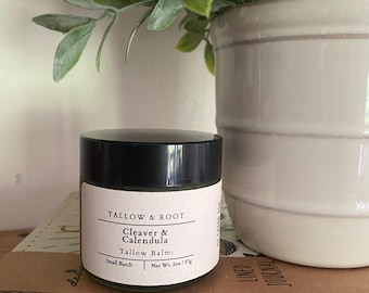 May include: A small glass jar with a black lid and a white label. The label reads "TALLOW & ROOT" and "Cleaver & Calendula Tallow Balm." The jar contains 2 oz / 57g of product. A green plant and a white ceramic pot are in the background.