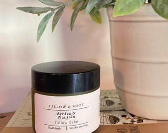 May include: A small, round jar of Tallow Balm with a black lid and a white label. The label reads "TALLOW & ROOT" and "Arnica & Plantain Tallow Balm." The jar is 2 oz / 57g. The jar sits on a book next to a white pot with green leaves.