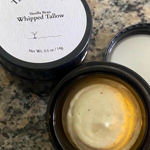 May include: A jar of Vanilla Bean Whipped Tallow with the lid off, revealing a creamy, off-white substance. The jar is black with a white label that reads "Tallow & Root" and "Whipped Tallow." The net weight is 0.5 oz / 14g.
