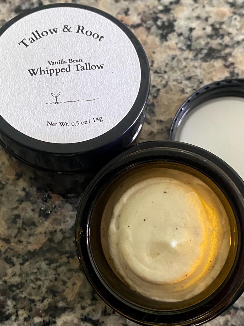 May include: A jar of Vanilla Bean Whipped Tallow with the lid off, revealing a creamy, off-white substance. The jar is black with a white label that reads "Tallow & Root" and "Whipped Tallow." The net weight is 0.5 oz / 14g.