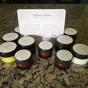 May include: A collection of small, round jars of Tallow & Root products, with black lids and white labels. The jars are arranged around a white card listing sample size products, including whipped tallow and balms. The background is a speckled countertop.