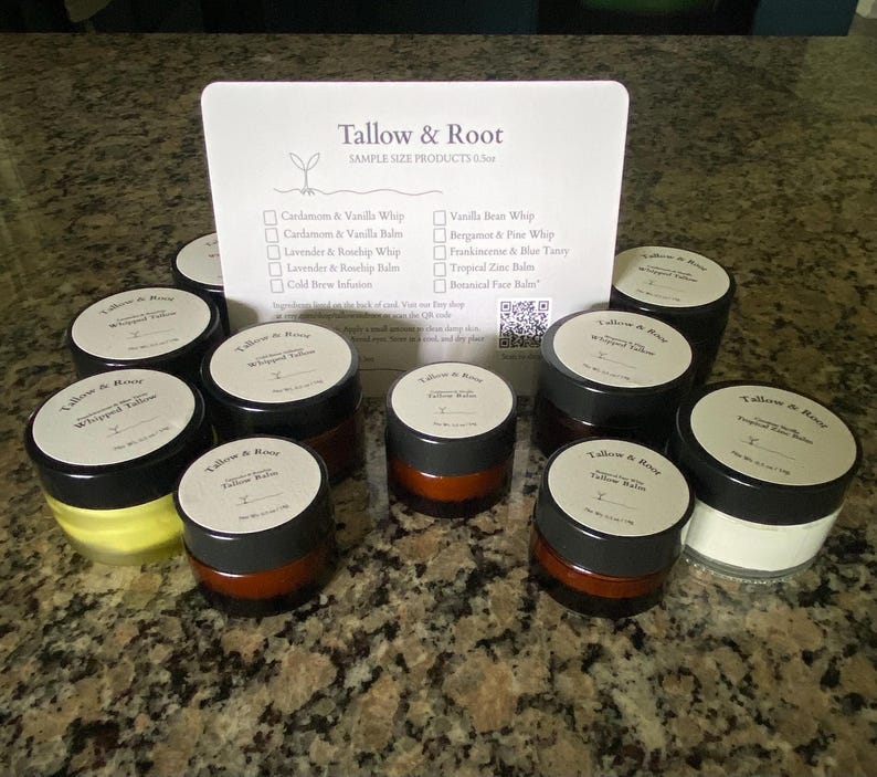 May include: A collection of small, round jars of Tallow & Root products, with black lids and white labels. The jars are arranged around a white card listing sample size products, including whipped tallow and balms. The background is a speckled countertop.