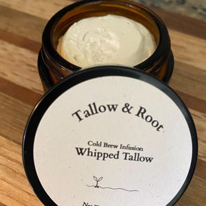 May include: An open amber glass jar filled with a creamy, off-white whipped tallow product. The lid, with the text "Tallow & Root" and "Whipped Tallow", is next to the jar. The net weight is 0.5 oz / 14g.