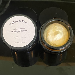 May include: Two small, black glass jars of whipped tallow. One jar is open, revealing a swirl of light yellow cream. The other jar is closed with a white label that reads "Tallow & Root" and "Cardamom & Vanilla Whipped Tallow". Net weight is 0.5 oz / 14g.