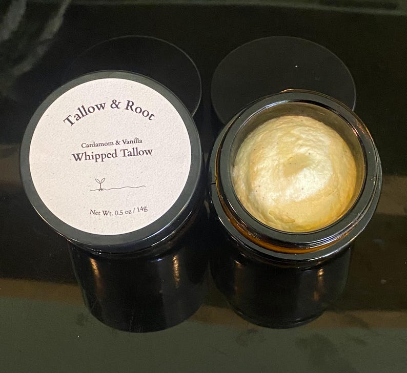 May include: Two small, black glass jars of whipped tallow. One jar is open, revealing a swirl of light yellow cream. The other jar is closed with a white label that reads "Tallow & Root" and "Cardamom & Vanilla Whipped Tallow". Net weight is 0.5 oz / 14g.