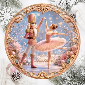 May include: A circular decorative plaque featuring a pink nutcracker and a ballerina in a pink tutu dancing together. The scene is framed by an ornate gold border with floral accents, set against a winter background with snowflakes and ornaments.