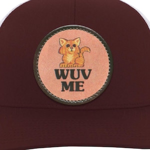 May include: A burgundy trucker hat with a white mesh back. The front features a circular patch with a cartoon cat and the words "WUV ME" in a bold font. The hat has a curved brim and a button on top.