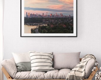 May include: A framed photograph of a cityscape at sunset hangs above a wooden sofa with pillows and a striped throw. A small wooden side table with a woven basket and decorative items sits on a jute rug.