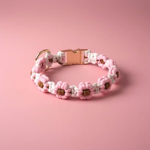 Puede incluir: Un collar para mascota de macramé con un patrón repetitivo de flores rosas y blancas con centros marrones. El collar está hecho de cuerda anudada e incluye una hebilla de liberación lateral de metal en oro rosa y una anilla en D a juego.